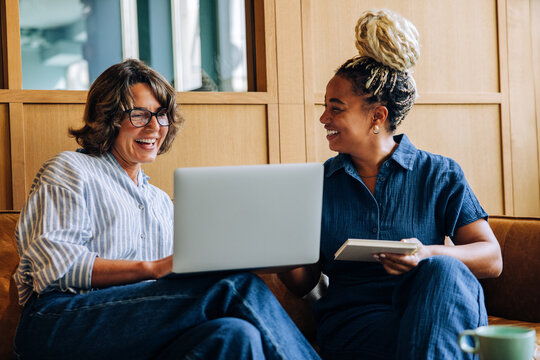 Two women smiling while using a laptop and notepad indoors