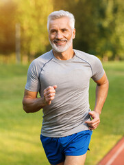 Fototapeta premium A man with gray hair runs happily on a track surrounded by green grass and trees. His athletic attire reflects his commitment to maintaining an active and healthy lifestyle.