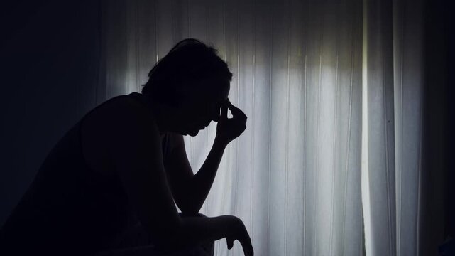 Silhouette of a woman sitting by a window, holding her head in distress, expressing anxiety, sadness, or emotional struggle. 4K.