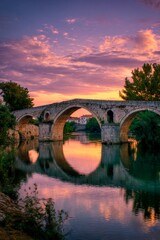 Fototapeta premium A serene sunset illuminates a historic stone bridge, creating stunning reflections in the tranquil river below. Lush greenery frames the scenic view