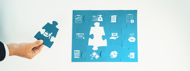 A hand is placing a puzzle piece into a blue puzzle featuring various financial symbols,...