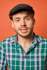 Close-up of smiling young Caucasian man wearing flat cap and green plaid shirt, orange background