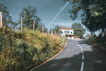Country road leading uphill to a rustic two-story house with blue shutters and weathered roof,...
