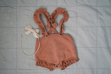 Knitted baby romper with ruffled straps and lace headband on quilted blanket.