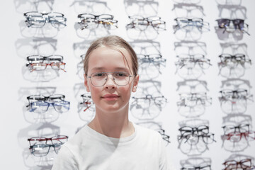 Child wearing eyeglasses stands inside an optical store with multiple frames displayed in the...