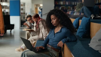 Focused biracial girl working on laptop studying in modern library with her diverse classmates discussing books in background - Powered by Adobe