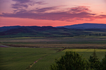 Sunset over rolling hills and agricultural lands. Scenic landscape featuring lush green agricultural fields in the foreground. Rolling hills under vibrant pink and purple sunset sky.