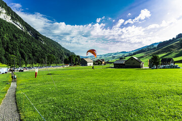 Scenic view of a paraglider flying above lush green alpine slopes and dramatic rocky peaks near...