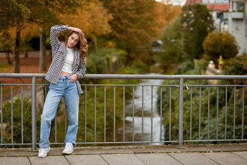 Young woman in trendy autumn outfit posing on a European bridge with golden foliage and a historic...