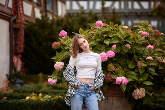 Stylish young woman posing in cozy autumn outfit among blooming hydrangeas in front of traditional half-timbered house in Germany, warm fall colors, outdoor portrait, casual fashion - Powered by Adobe
