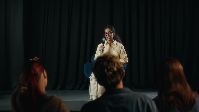 Full shot of young biracial female author sitting on chair on stage and interacting with young audience in warm and friendly manner during book presentation or seminar