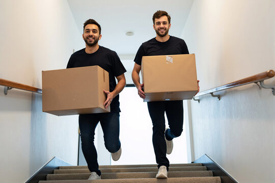 Two happy male movers run up a staircase while carrying cardboard boxes, providing a fast, energetic, and efficient relocation and delivery service with a smile