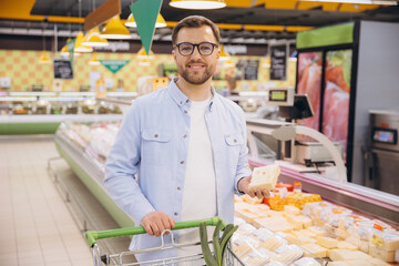 Man choosing cheese in refrigerated section of supermarket pushing shopping cart