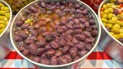 A close-up view of black or dark purple olives, immersed in a brine, inside a metal container. The...