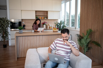 Focused freelancer working from home while partner reads in kitchen