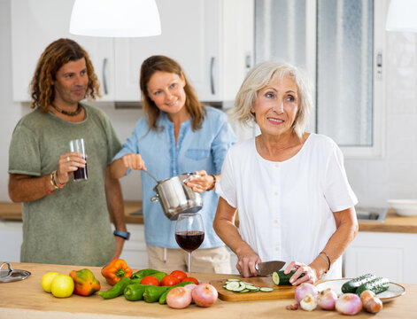 Joyful old female cutting vegetables on board during the rest of man and women with wine at the modern cuisine