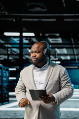 Admin listening music in data center, using tablet to oversee infrastructure mainframes. Worker...