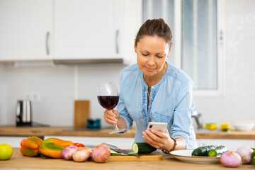 Portrait of young adult woman holding glass of wine and browsing on her smartphone, standing near...