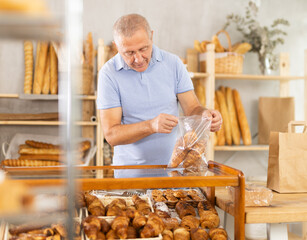 Senior man in bakery is picking up buns for tea. Client uses tongs to take croissants out of glass...