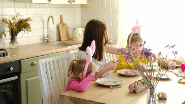 Children siblings painting Easter egg sitting together at table. Easter Family traditions. Teen girl teaching happy little brother and sister to dye, decorate eggs with paints. Preparation to holidays