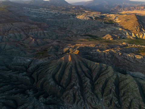 A striking aerial view of rugged hills and deep valleys shaped by natural erosion, revealing dramatic textures and earthy tones under the warm, directional light of the early morning sun.