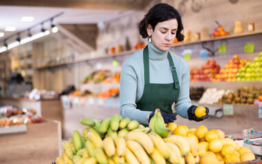 While working in shop, employee forms display case, puts ripe imported lemon in pile. Open display of goods in self-service shop. Female employee makes attractive display case with vegetables.