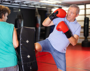 Elderly man practices kicking and punching with a trainer in a gym