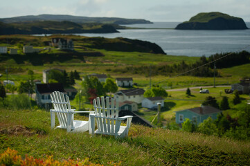 White Adirondack chairs overlook rolling grassy hills that slope to calm coastal waters, framed by...