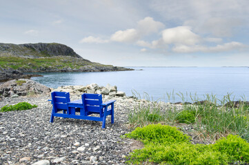 A blue Adirondack bench seat rests on a rocky shoreline in Fogo, Fogo Island, overlooking the calm...