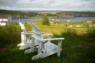 White Adirondack chairs on a grassy bluff offer expansive ocean views where waves meet rocky shore,...