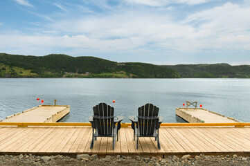 Two dark Adirondack chairs sit side by side on a wooden dock in Trinity, Newfoundland, facing the...