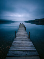 Obraz premium A tranquil lakeshore scene with a long wooden dock, still waters, and soft mist rising at sunrise. Photorealistic rendering emphasizes natural beauty and the calm serenity of early morning.