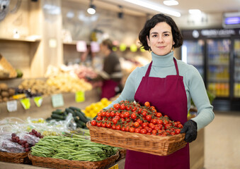 Woman in apron carries large quantity of tomato in wicker basket to sales area, supplementing stores assortment with fresh vegetables