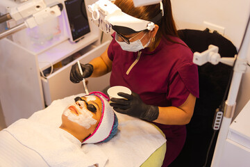Beautician applies a facial mask with a brush to a reclining client, wearing a magnifying visor in...