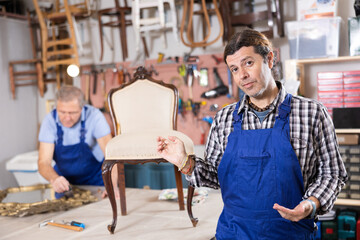 Professional middle-aged craftsman restorer in uniform renewing antique chair while working in repair shop