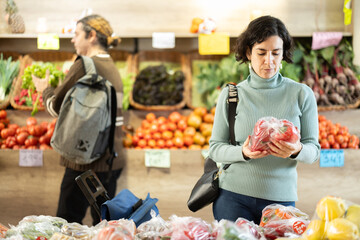 European woman, standing near the vegetable counter, examines and chooses a bell pepper packed in a plastic bag. Buyer is preparing to weigh the peppers before buying