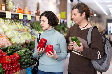 European couple stands in a store near a vegetable counter and chooses fresh cucumbers and red bell...