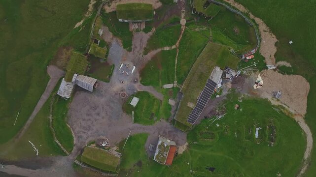 Drone captures a slow, steady view of a circular Icelandic turf house settlement, with grass roofs, pathways, and solar panels in lush green fields.