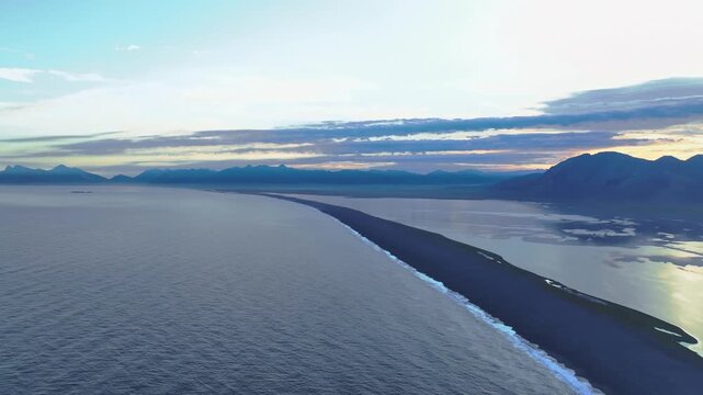 Sweeping aerial view of a black sand beach bordered by ocean and water bodies, with rugged mountains on the horizon under a soft blue and orange sky.