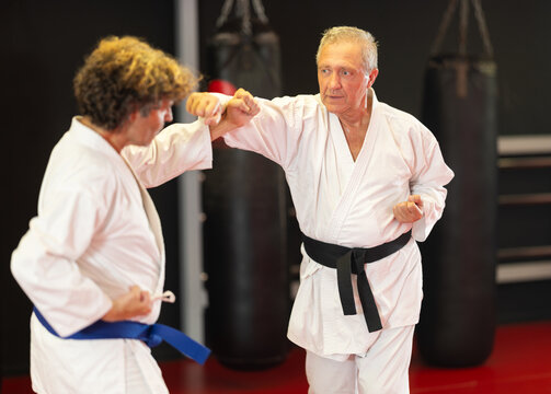 Focused older karate fighter in white gi with black belt practicing paired kumite, honing punch technique with partner during training in martial arts dojo with red mats and hanging punching bags.. - Powered by Adobe