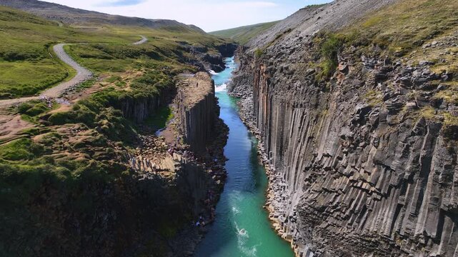 Aerial view of Studlagil Canyon in Iceland, showing basalt columns, a turquoise river in motion, visitors on rocky edges, and lush green hills.