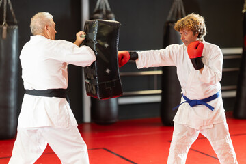 Determined blue-belt fighter wearing red gloves punching padded shield under watchful guidance of experienced black-belt partner in modern martial arts dojo..