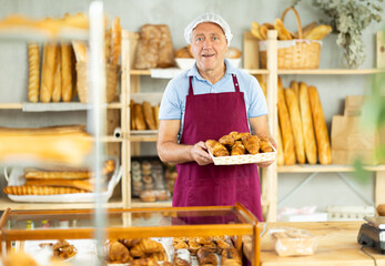 Elderly male salesperson in apron holding basket with croissants in bakery