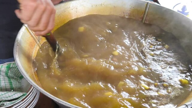 
A person stirs a large pot of a thick, yellow-green mixture, a traditional Indonesian soto ayam soup, then scoops it out with a ladle to serve.