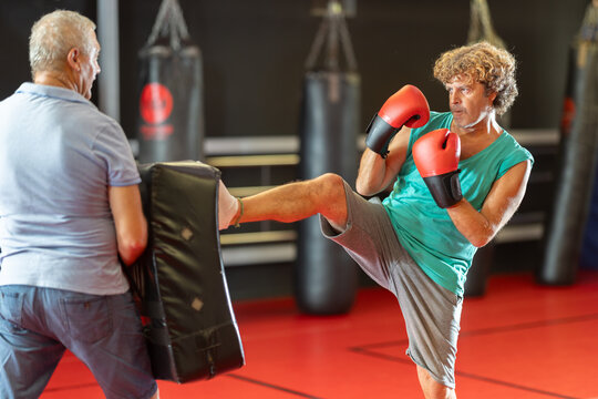 Determined middle-aged hobby kickboxer driving solid kick into partners shield during practical self-defense drill on red mats at local gym