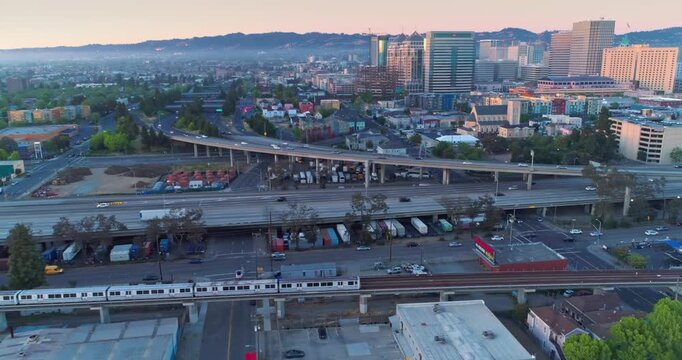 Aerial view of Oakland showing the city skyline, highway traffic, and a BART train crossing over the freeway during the early morning hours.  Oakland, California, USA. 11 May 2025.