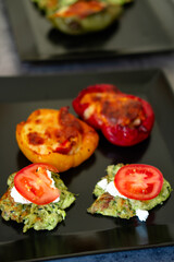 Stuffed bell peppers and zucchini fritters with tomato topping on black plates.