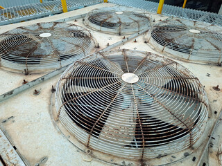 A weathered industrial air conditioner unit with multiple large fan blades, showing signs of rust and age on a rooftop. The metallic surface and grates are in need of maintenance