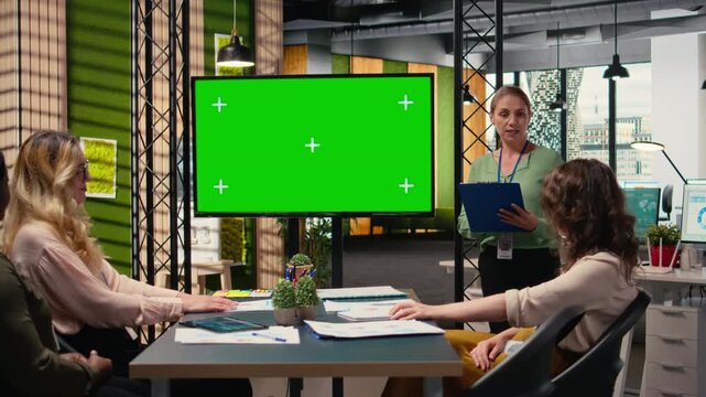 Multiethnic corporate staff gathers in front of a green screen TV display, preparing a pitch that will feature financial data reports and graphs for a strategy development plan. Camera A.