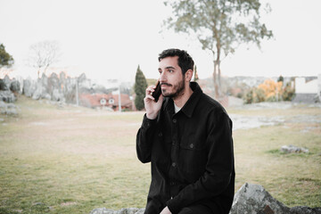 Confident young Hispanic man using a smartphone in the park, wearing a black jacket